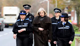 Theresa May, looking like a cross (cross!) between Cruella de Ville and an avenging angel, accompanied by a goon-squad of police.