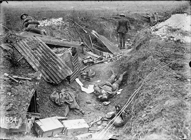 A German machine gun position photographed just after its capture by New Zealand troops during World War I. Shows a New Zealander inspecting the trench in which a dead German soldier lays. Sheets of iron and pieces  of timber are strewn around the nearby dugout. Two stick hand grenades  are visible in the foreground. Photograph taken Grevillers 24 August  1918 by Henry Armytage Sanders