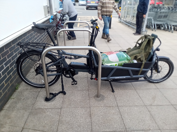 A grey cargo bike locked to a bicycle stand outside a building, with a carpark behind