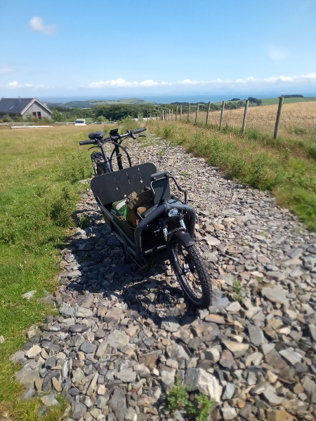 A grey cargo bike parked in sunlight on a green hilltop, looking out and down towards the sea far below