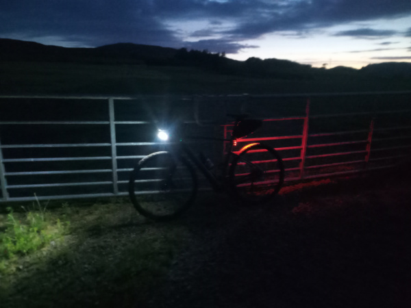 My Cannondale Topstone Neo, leaning against a gate towards the end of it's first off-road nighttime adventure. A dark grey bike with bright lights, leaning against a grey field gate, with the landscape behind lost in darkness. Above the outline of the hills, the sky retains some brightness