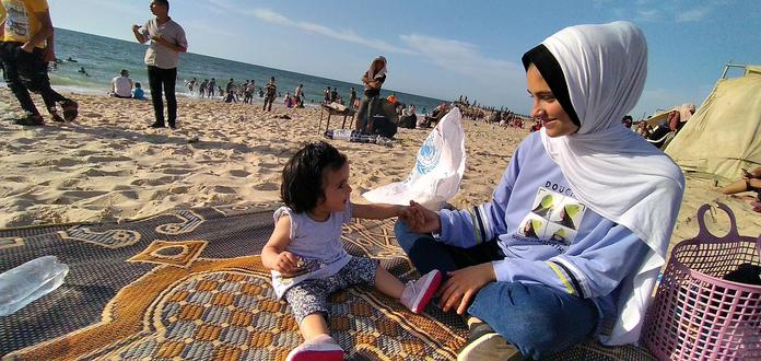 Aya, a teenager in a blue sweatshirt and a headscarf, and Kinda, a small child, sitting on a rug on a sandy beach.
