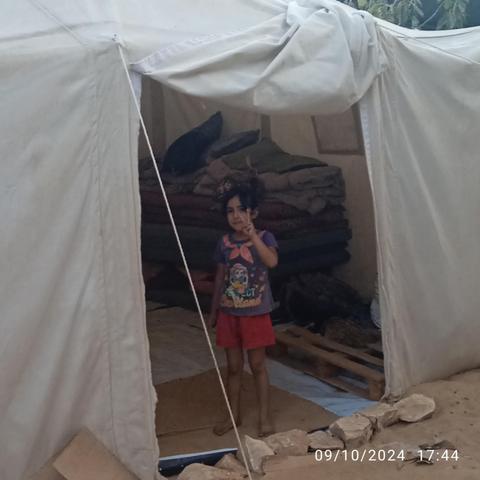 A small child stands in the doorway of a square white tent. There are mats on the floor inside.