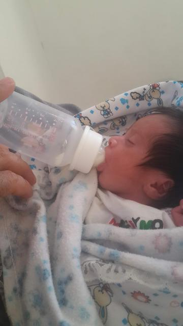 A tiny baby, swaddled in a patterned white blanket, drinking milk from a bottle that seems disproportionately large.