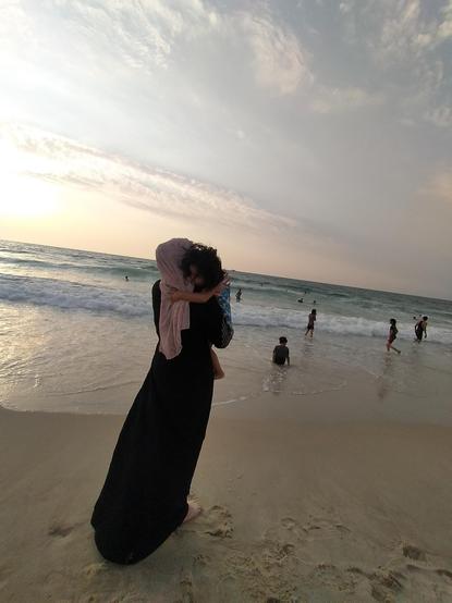 Aya carries her sister Kinda along the beach, in evening light.