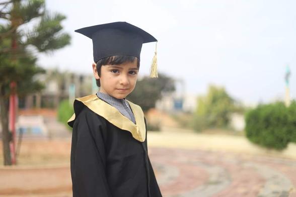 Ady, a young boy proudly wearing full academic robes, stands in a sandy yard with bushes and trees behind.