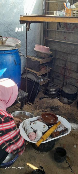 A woman in a headscarf sits on the bare sand, washing up in a small basin, with a tray of washed crockery beside her. In the background is a pile of cooking pots.