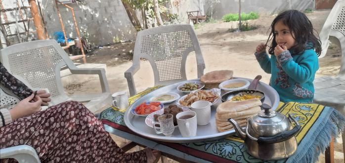 A small child (Kinda?) in a blue jacket, beside an outdoor table on which is a large circular tray with many dishes of food