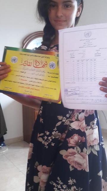 Aya, a teenage girl, holding a brightly coloured certificate and a typed sheet of white paper showing scores. She looks serious, but pleased.