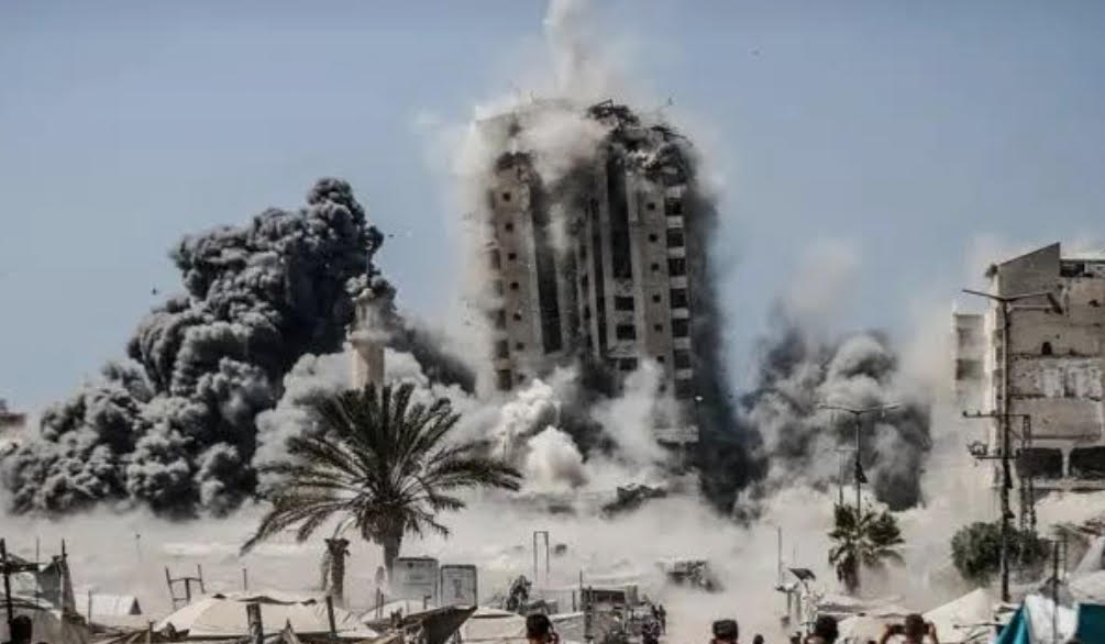 A multi-story apartment building collapses, among other bombed-out buildings