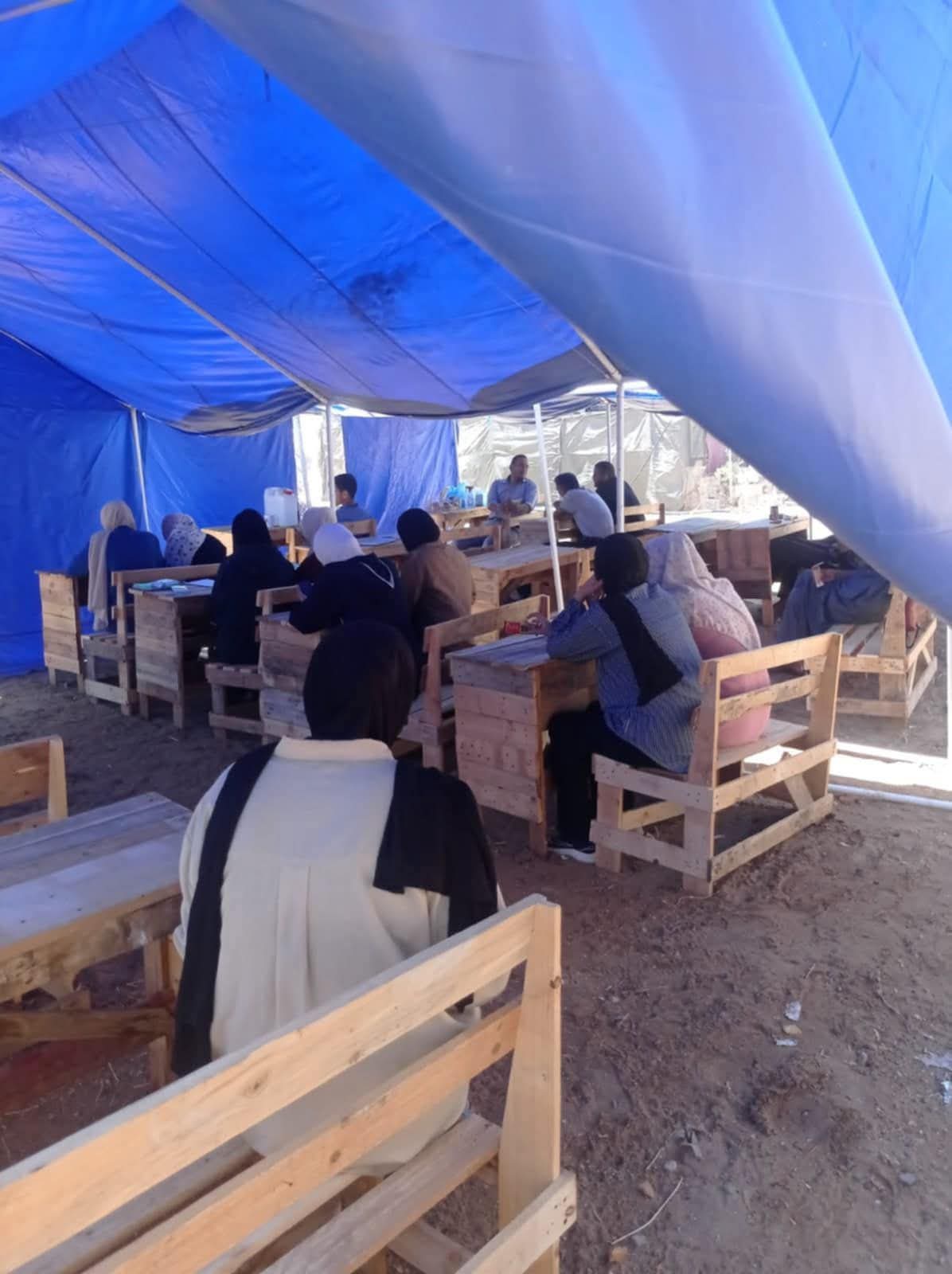 A very large makeshift tent, set out as a classroom, with crudely made desks and chairs